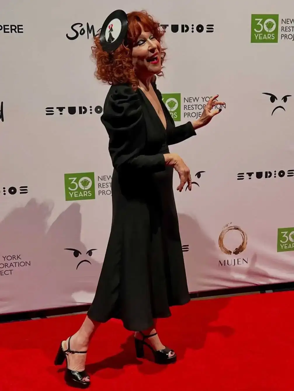 Singer and actress Bette Midler poses on a red carpet in a black long-sleeved dress and a whimsical vinyl record headpiece.